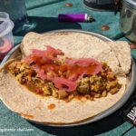 Shown is a serving of pork tacos with Mexican rice, arranged on a soft tortilla wrap. This recipe features dehydrated ingredients all made at home, such as pork, rice, tomatoes, onions and carrots. We’ll rehydrate the dry mix and use it as a tortilla wrap filling. Optionally, we added some pickled onions and topped it off with salsa.